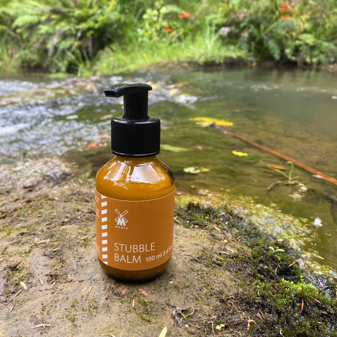 MÜHLE Stubble Balm in a glass jar placed on a riverbank rock with flowing water and natural outdoor surroundings