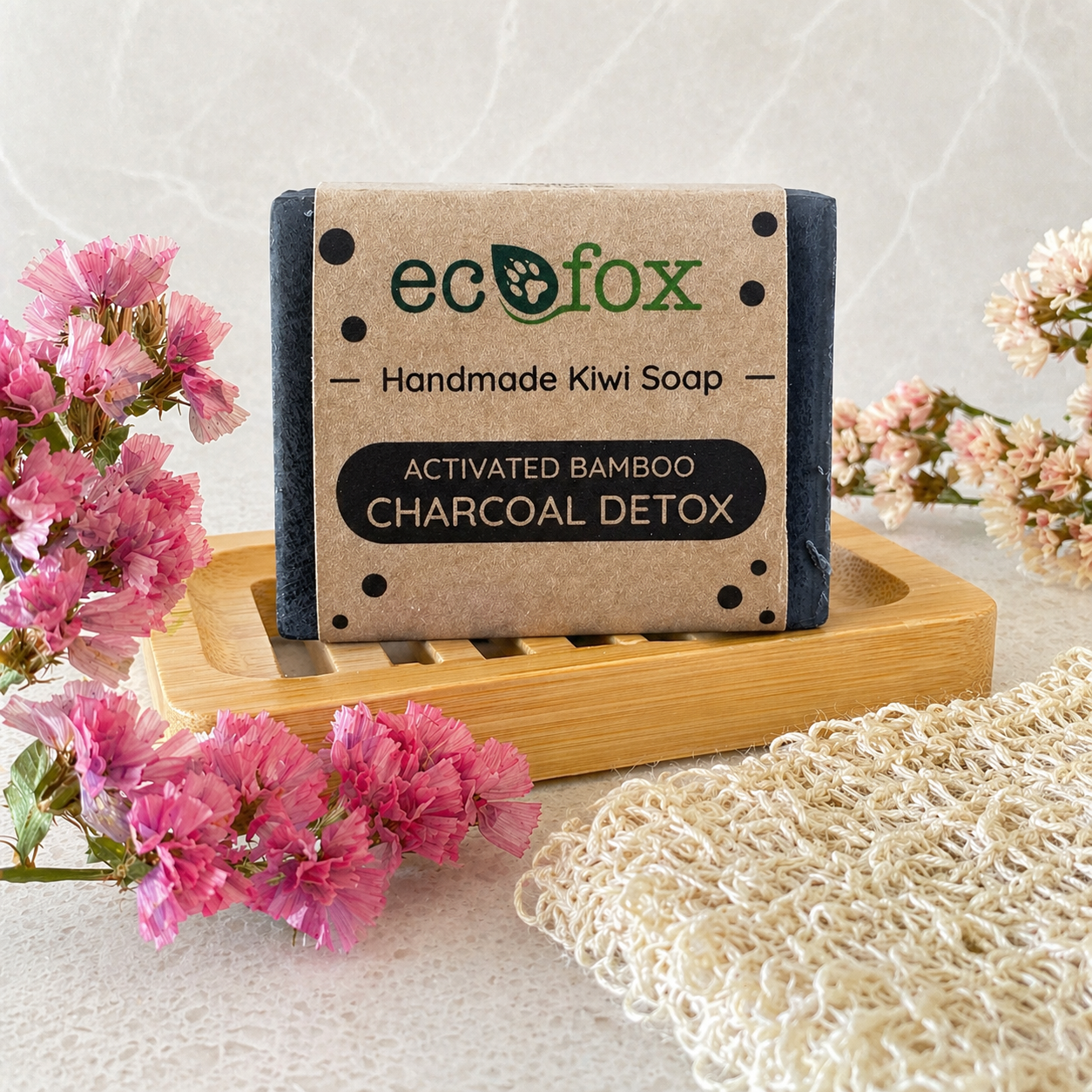 Eco Fox Activated Bamboo Charcoal Detox body soap displayed on a bamboo soap dish in a soft spa-style bathroom setting with pink flowers and natural textured cloth.