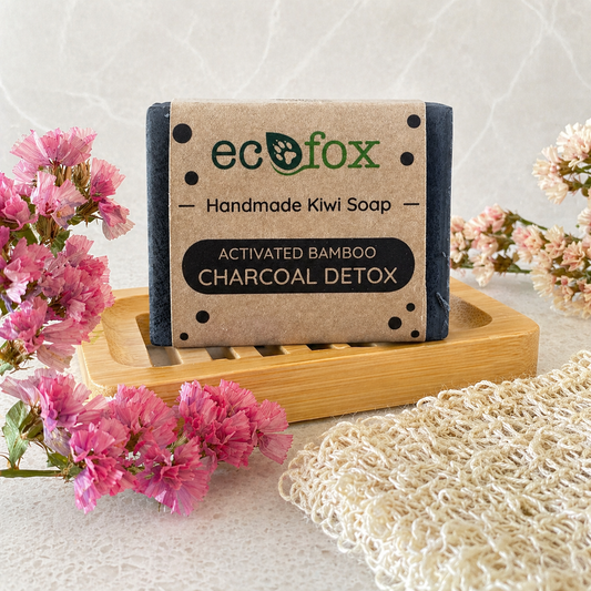Eco Fox Activated Bamboo Charcoal Detox body soap displayed on a bamboo soap dish in a soft spa-style bathroom setting with pink flowers and natural textured cloth.