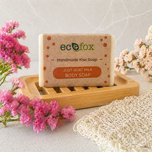 Eco Fox Just Goat Milk body soap displayed on a bamboo soap dish in a soft spa-style bathroom setting with pink flowers and natural textured cloth.